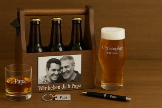 Personalisierte Vatertagsgeschenke auf Holztisch: gravierter Beer Caddy mit Foto, Bierglas mit Name, Whiskyglas, Schlüsselanhänger und Kugelschreiber mit „Papa“-Gravur – ideal für Vatertag 2025.