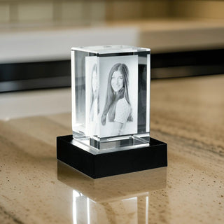 Der 2D-Glasblock Hochformat von Engravy zeigt eine lächelnde junge Frau mit langem Haar aus Kristallglas, die auf einem schwarzen Sockel ruht und elegant auf einer gemusterten Tischplatte präsentiert wird.