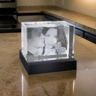 Der Engravy 2D-Glasblock Querformat – Individuelle Gravur aus Kristall schmückt die Küchentheke mit einem eingravierten Bild eines Paares. Der Mann küsst die Frau und sie posiert für die Kamera. Es ist auf einem LED-Leuchtsockel montiert, der einen blendenden reflektierenden Glanz erzeugt.
