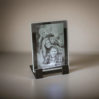 Ein Graustufenfoto von vier lächelnden Menschen, zwei Männern und zwei Frauen, ist elegant in einem eleganten Kristallglas von Engravy gerahmt. Die 2D-Glasscheibe im Hochformat sitzt auf einem schwarzen Ständer und wird anmutig auf einer glatten, hellen Oberfläche präsentiert.