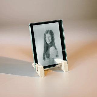 Ein eingraviertes monochromes Foto einer lächelnden Person mit langen Haaren und hellem Oberteil wird auf einer Engravy 2D-Glasscheibe im Hochformat angezeigt, elegant gerahmt in Kristallglas. Es wird von einem Holzständer auf einer neutralen Oberfläche getragen und verleiht jeder Umgebung zeitlosen Charme.