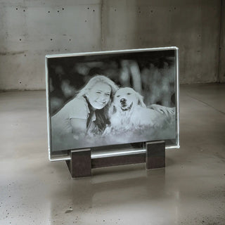 Engravys 2D-Glasscheibe im Querformat zeigt das eingravierte Bild einer lächelnden Person mit einem glücklichen Hund im Freien, umgeben von Gras, auf einem Metallständer. Die Szene wird durch einen LED-Leuchtsockel vor Betonwänden und -böden verstärkt.