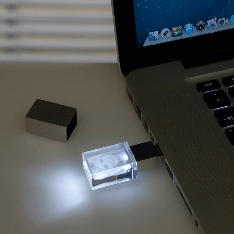 Ein Engravy 2D-USB-Stick mit Foto-Gravur, der ein leuchtendes Kristallglas-Element mit einem Gesicht aufweist, ist an einen Laptop angeschlossen. Der Bildschirm ist mit Symbolen gefüllt und die USB-Abdeckung liegt daneben auf dem Tisch.