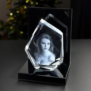 Der Rahmen Engravy 3D-Glasgravur Diamond im eleganten Premiumformat zeigt das Porträt einer langhaarigen Frau auf schwarzem Sockel. Dieser unregelmäßig geometrische Kristall fängt Eleganz inmitten verschwommenen grünen und goldenen Lichts ein.