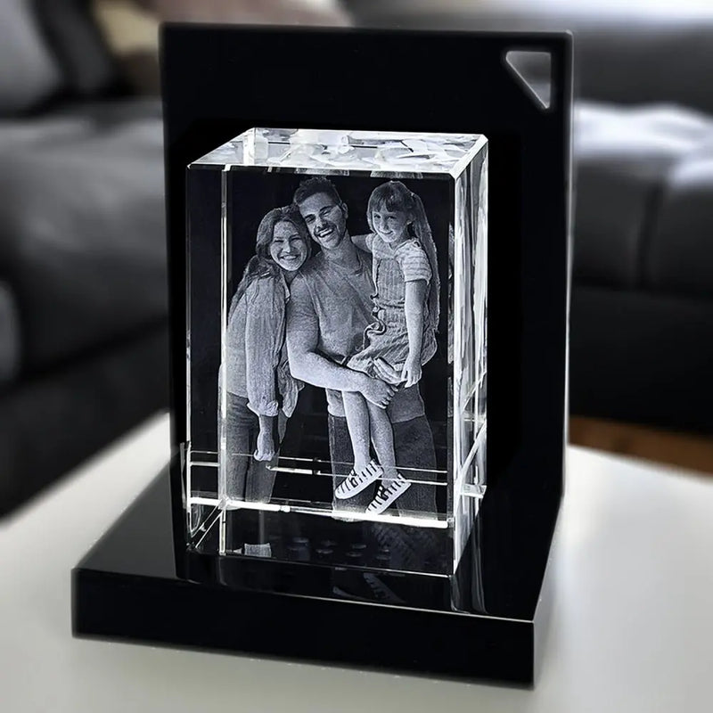 Der Engravy 3D-Glasblock Hochformat zeigt eine detaillierte 3D-Gravur einer lächelnden Familie, die in einen Kristallwürfel eingraviert ist. Er sitzt auf einem LED-Leuchtsockel vor einem verschwommenen Wohnzimmer und beleuchtet das komplizierte Design.