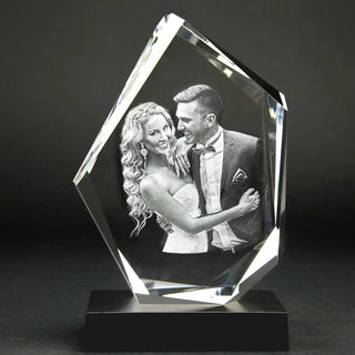 Eine atemberaubende 3D-Glasgravur eines lächelnden Paares in formeller Kleidung. Der Mann im Anzug und die Frau im eleganten Kleid umarmen sich liebevoll. Dieses exquisite Kristallglasstück hat eine unregelmäßige geometrische Form und ruht elegant auf einem schwarzen Sockel. Produkt: Der Diamant | Elegantes Premium-Format.