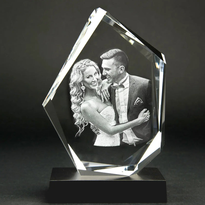 Eine atemberaubende 3D-Glasgravur eines lächelnden Paares in formeller Kleidung. Der Mann im Anzug und die Frau im eleganten Kleid umarmen sich liebevoll. Dieses exquisite Kristallglasstück hat eine unregelmäßige geometrische Form und ruht elegant auf einem schwarzen Sockel. Produkt: Der Diamant | Elegantes Premium-Format.