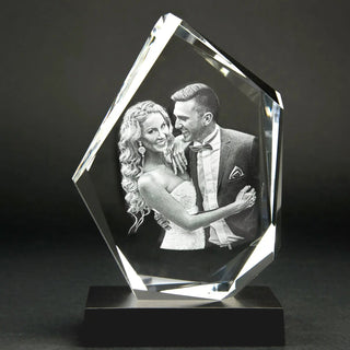 Eine atemberaubende 3D-Glasgravur eines lächelnden Paares in formeller Kleidung. Der Mann im Anzug und die Frau im eleganten Kleid umarmen sich liebevoll. Dieses exquisite Kristallglasstück hat eine unregelmäßige geometrische Form und ruht elegant auf einem schwarzen Sockel. Produkt: Der Diamant | Elegantes Premium-Format.