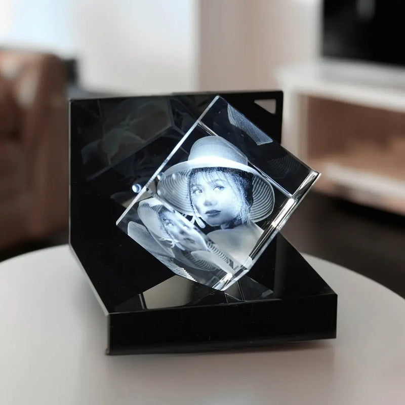 Ein Engravy 3D-Glaswürfel mit Gravur ruht elegant auf einem LED-Leuchtsockel, der auf einem runden Tisch vor einem sanft verschwommenen Wohnzimmerhintergrund angeordnet ist und so Tiefe und Faszination verleiht.