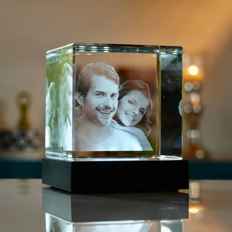 Ein Engravy 3D-Glaswürfel – ein personalisiertes Meisterwerk aus Kristall mit 3D-Gravurporträts eines lächelnden Mannes und einer lächelnden Frau sitzt auf einem reflektierenden Tisch. Die sanfte Unschärfe und die warme Beleuchtung verstärken die gemütliche Atmosphäre.