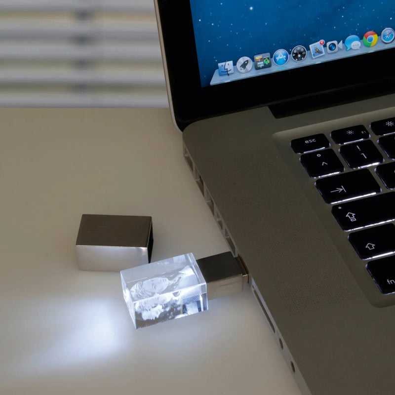 Ein Laptop mit eingestecktem Engravy 3D-USB-Stick mit Lasergravur, dessen transparentes Gehäuse und filigranes Design strahlend weiß leuchten, bietet 16 GB Speicherkapazität. Der Bildschirm zeigt Desktopsymbole auf hellem Hintergrund.