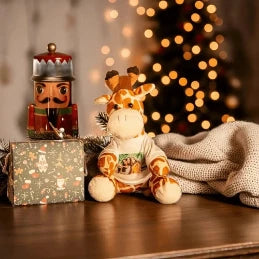 Personalisiertes Plüschtier einer Giraffe mit einem bedruckten T-Shirt, sitzt vor einem Nussknacker und einem verpackten Weihnachtsgeschenk auf einem Holztisch. Im unscharfen Hintergrund ist ein weihnachtlich beleuchteter Baum zu sehen, was eine festliche Weihnachtsatmosphäre schafft. Ideal als individuelles Fotogeschenk für Weihnachten.