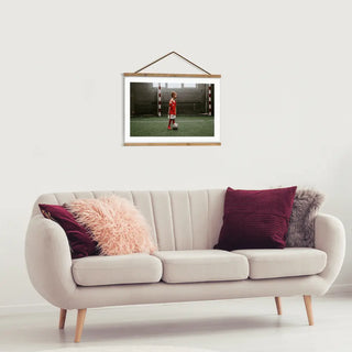 Das Clipwood Wandbild mit Magnetleisten von Engravy, das einen Fußballspieler in Rot und Weiß zeigt, ist über einem cremefarbenen Sofa mit Holzbeinen angebracht. Mit rosa und burgunderfarbenen Kissen geschmückt, vermittelt die Kulisse eine schlichte, moderne Wohnzimmeratmosphäre mit minimalistischem Charme.