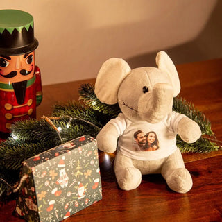 Ein Engravy Kuschelelefant mit Foto – Dein personalisiertes Stofftier ruht auf einem Holztisch, der mit festlichen Dekorationen wie einem Nussknacker, einem verpackten Geschenk und beleuchteten immergrünen Zweigen geschmückt ist. Dieser personalisierte Plüschelefant dient als ideales individuelles Geschenk für jeden Anlass.