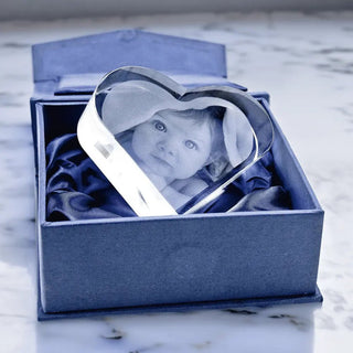 Ein graviertes Liegendes 2D-Glasherz – Personalisiertes Fotogeschenk mit einem Kinderfoto wird elegant in einer offenen blauen Samtbox präsentiert. Die 2D-Fotogravur zeigt den sanften Ausdruck des Kindes in einem hellen Gewand, das auf einer Marmoroberfläche ruht.