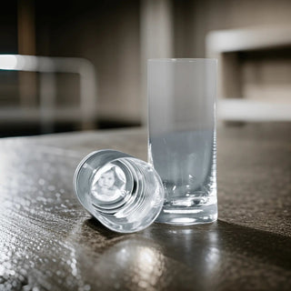 Zwei Engravy Long-Drink-Gläser stehen auf einem Holztisch, eines aufrecht und das andere flach liegend, und spiegeln ein verzerrtes Schwarz-Weiß-Bild wider. Diese personalisierbaren Gläser mit Lasergravur eignen sich ideal als Geschenk für besondere Anlässe. Der sanft verschwommene Hintergrund sorgt für eine elegante Note.