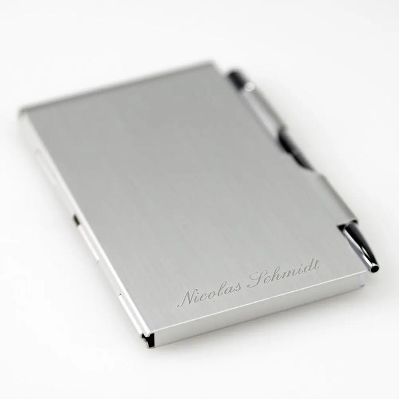 Der Notizblock mit Gravur von Engravy ist ein eleganter Visitenkartenhalter aus Aluminium mit minimalistischem Design und integriertem Stift. Er trägt den eingravierten Namen „Nicolas Schmidt“ und verleiht dem schlichten weißen Hintergrund eine elegante Note, perfekt für jeden Berufstätigen.