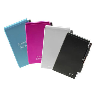 Der „Notizblock mit Gravur – Praktisch & elegant“ von Engravy umfasst vier geschlossene Notizbücher in leuchtendem Blau, Pink, Silber und Schwarz, jeweils mit integriertem Stift. Sie sind mit Namen und Initialen in weißer Schrift geprägt und weisen ein Design auf, das an hochwertige Aluminiumetuis erinnert.