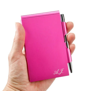 Eine Hand hält einen leuchtend rosa Engravy-Notizblock mit Gravur – praktisch & elegant, an dessen rechter Seite ein Drehkugelschreiber angebracht ist. „A.J.“ ist elegant in die untere rechte Ecke eingraviert und hebt sich von einem schlichten weißen Hintergrund ab.