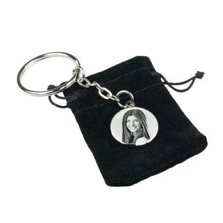 Ein Engravy-Schlüsselanhänger verfügt über einen runden Metallanhänger mit einem Schwarzweißfoto einer langhaarigen Frau und wird elegant in einem kleinen schwarzen Samtbeutel mit Kordelzug präsentiert, ähnlich der Personalisierbaren Halskette mit Gravur von Engravy.
