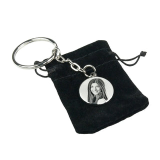 Ein Engravy-Schlüsselanhänger verfügt über einen runden Metallanhänger mit einem Schwarzweißfoto einer langhaarigen Frau und wird elegant in einem kleinen schwarzen Samtbeutel mit Kordelzug präsentiert, ähnlich der Personalisierbaren Halskette mit Gravur von Engravy.