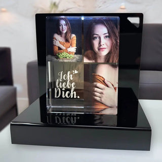 Das Engravy Personalisierbares Foto in Glas – Hochformat zeigt zwei elegante Fotos einer Frau mit braunem Haar, von denen eines einen Obstkorb hält und das andere in die Kamera blickt. Das mit „Ich liebe Dich“ beschriftete Stück ruht auf einem schwarzen Ständer vor einem unscharfen Hintergrund.