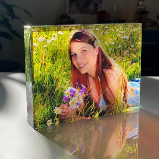 Ein personalisierbares Glasfoto im Querformat von Engravy zeigt eine lächelnde Person mit langen Haaren, die zwischen Wildblumen liegt und Gänseblümchen und lila Blüten hält. Das Sonnenlicht wird vom Block reflektiert und macht ihn zu einem zeitlosen Erinnerungsstück.