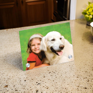 Ein personalisierbares Glasfoto mit Alufüßen von Engravy steht elegant auf einer Arbeitsplatte aus Granit und zeigt das Bild eines Kindes in einem roten Hemd, das lächelt und einen großen weißen Hund umarmt, umgeben von grünem Gras.