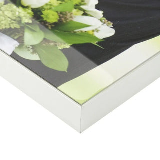 Eine Nahaufnahme einer Ecke zeigt ein gerahmtes Bild mit Blumenmuster, das einem exquisiten Fotodruck ähnelt. Der weiße Rahmen umschließt grüne und weiße Blumen und hebt die glänzende Oberfläche hervor. Elegant und stilvoll wie Engravys personalisierte Glas-Wanduhr.