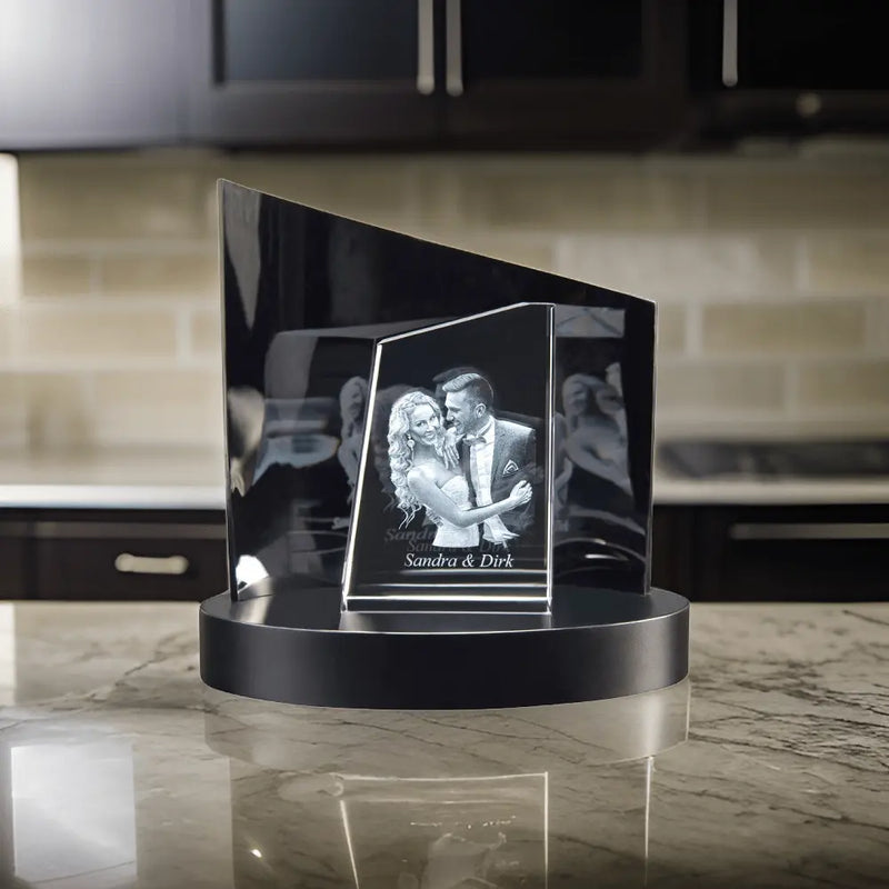 Engravys Premium 3D-Glasgravur – Der Turm zeigt ein tanzendes Paar, das elegant auf einem schwarzen runden Sockel auf einer Marmorarbeitsplatte balanciert. Im verschwommenen Küchenhintergrund steht auf der Gravur „Besonderer Moment Sandra & Dirk“, was das außergewöhnliche Design für besondere Anlässe hervorhebt.
