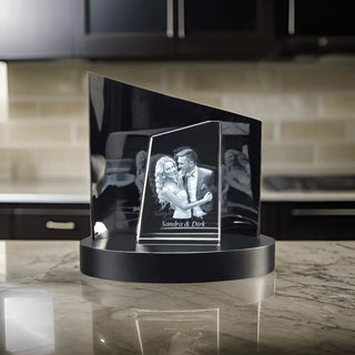 Engravys Premium 3D-Glasgravur – Der Turm zeigt ein tanzendes Paar, das elegant auf einem schwarzen runden Sockel auf einer Marmorarbeitsplatte balanciert. Im verschwommenen Küchenhintergrund steht auf der Gravur „Besonderer Moment Sandra & Dirk“, was das außergewöhnliche Design für besondere Anlässe hervorhebt.