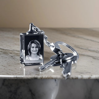 Ein Engravy-Schlüsselanhänger mit 2D-Gravur – Verschiedene Formen, bestehend aus zwei silbernen Schlüsseln und einem rechteckigen Kristallblock mit eingravierter lächelnder Person, ruht elegant auf einer Marmoroberfläche mit sanft verschwommenem Hintergrund – ideal für jede Geschenkbox.