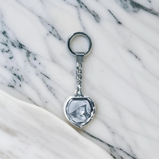 Ein herzförmiger silberner Schlüsselanhänger mit 2D-Gravur von Engravy liegt auf Marmor. Er verfügt über einen transparenten Abschnitt mit einem Katzenfoto und ist somit ein perfektes Geschenk für Tierliebhaber. Hübsch verpackt in einer bezaubernden Geschenkbox ist er ein ideales Andenken.