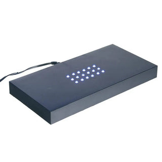 Der Engravy Sockel, ein schwarzes rechteckiges Gerät mit glatter Oberfläche, ähnelt einem modernen Glasblock. Auf der Oberseite befinden sich kleine blaue LEDs in einem Gittermuster, die sein schlankes Design unterstreichen. Für die Verbindung ist ein schwarzes Kabel angeschlossen.