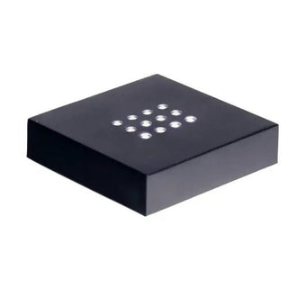 Die Gravur „Sockel Scheibe selbstehend Batteriefach“ ist eine schwarze, rechteckige Box mit einem Gitter aus kleinen, weißen, runden Lichtern auf der Oberseite, die an die Ästhetik von Glasblock erinnert. Von der Seite betrachtet betont das flache Design die Eleganz der Anordnung.