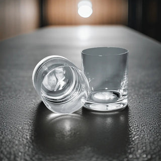 Zwei Engravy Trinkgläser mit Gravur (400 ml) stehen auf einer dunklen Tischplatte. Eines steht aufrecht, das andere liegt auf der Seite und gibt den Blick auf ein Schwarz-Weiß-Foto im Inneren frei. Der sanft verschwommene Hintergrund suggeriert eine helle Lichtquelle.