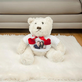 Der Weiße Teddybär mit Foto – Persönliches Kuscheltier mit Herz von Engravy sitzt auf einem weißen, flauschigen Teppich, trägt ein T-Shirt mit dem Foto eines lächelnden Paares und ist um den Hals mit zwei roten Herzen geschmückt, vor dem Hintergrund eines gemütlichen beigen Sofas.